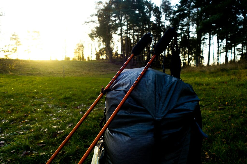 A black backpack with orange trekking poles rests on green grass, framed by a sunrise and trees in the background.
