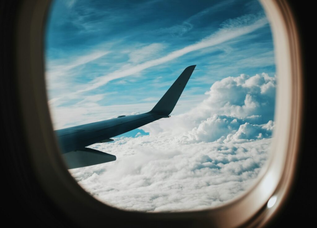 View from an airplane window showcasing a wing above a sea of fluffy clouds against a bright blue sky with wispy clouds.