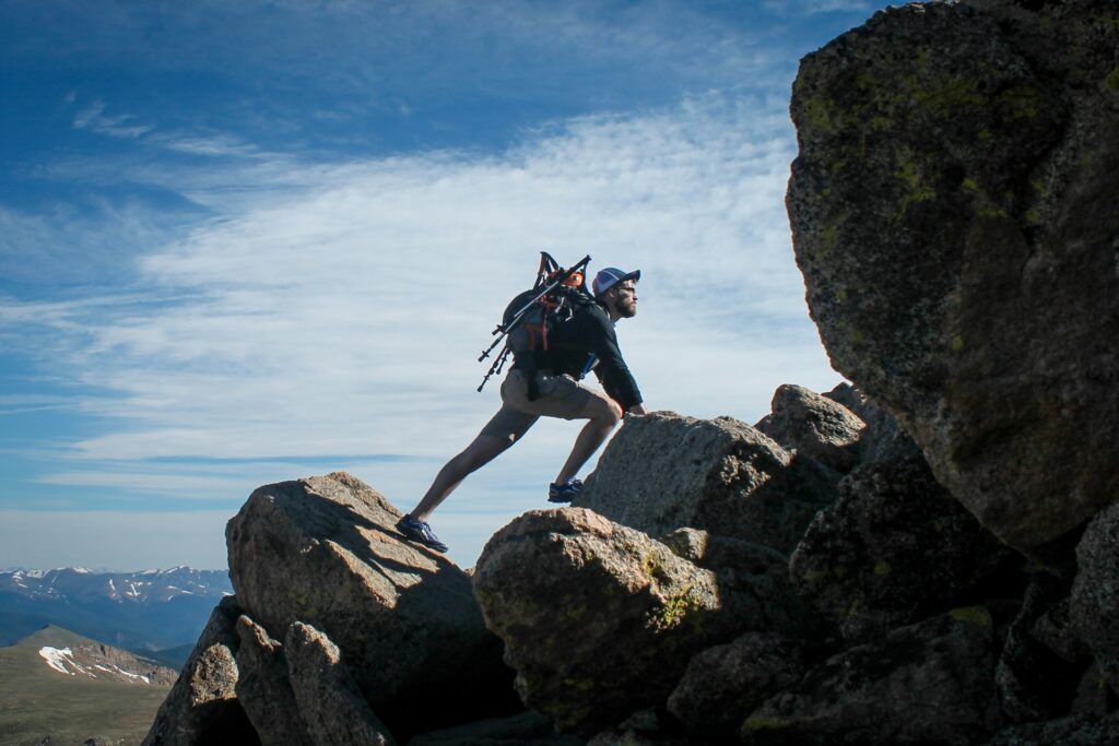 A hiker climbs over a rocky terrain against a backdrop of mountains and blue sky, showcasing determination and adventure.