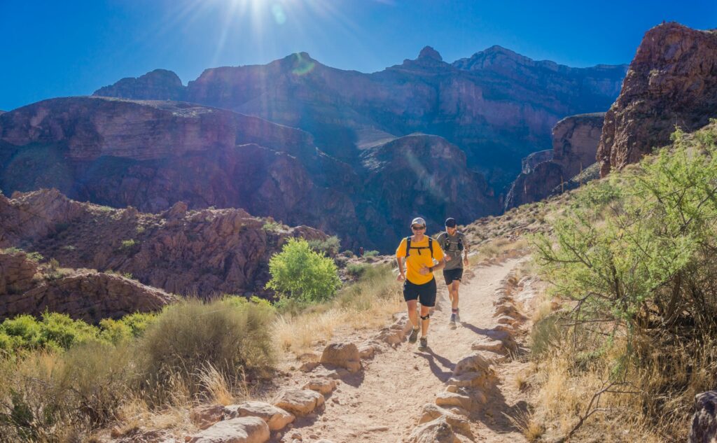 Two hikers traverse a sunlit trail through rocky terrain, surrounded by steep canyon walls and vibrant greenery.