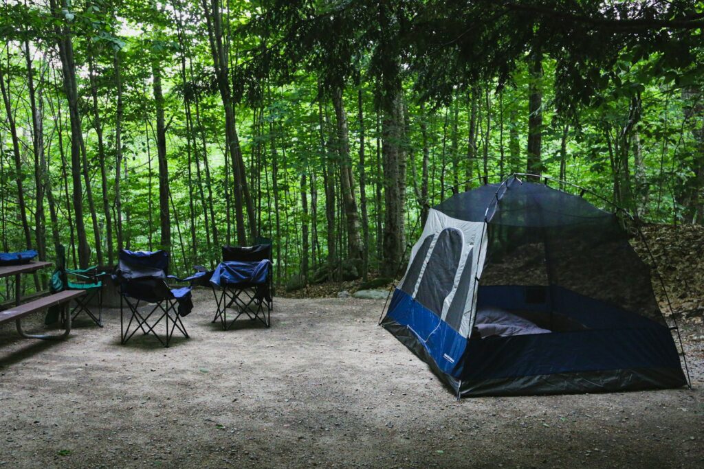 A cozy camping setup features a tent and blue folding chairs surrounded by lush green trees, creating a serene outdoor atmosphere.