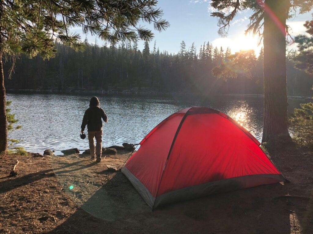 A camper strolls by a serene lake at sunrise, with a red tent nestled among trees, reflecting nature's tranquility.