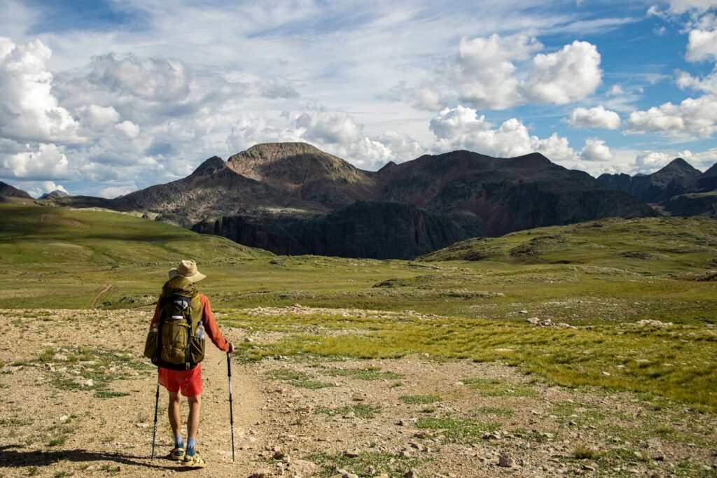 A hiker in a wide landscape, wearing a backpack and using trekking poles, gazes at distant mountains under a cloudy sky.