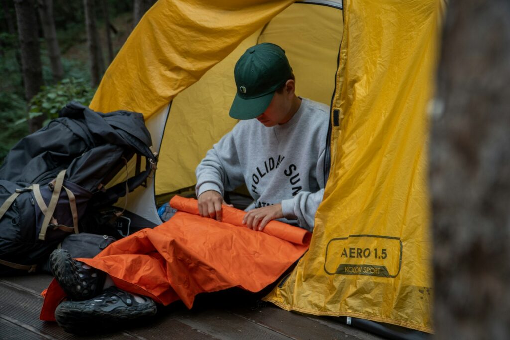 A man sits inside a tent, next to an orange bag, surrounded by camping gear and a cozy atmosphere.