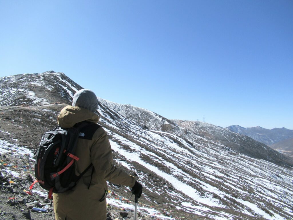 A hiker in a brown jacket and gray beanie stands on a snowy mountain, gazing at the distant peaks under a clear blue sky.
