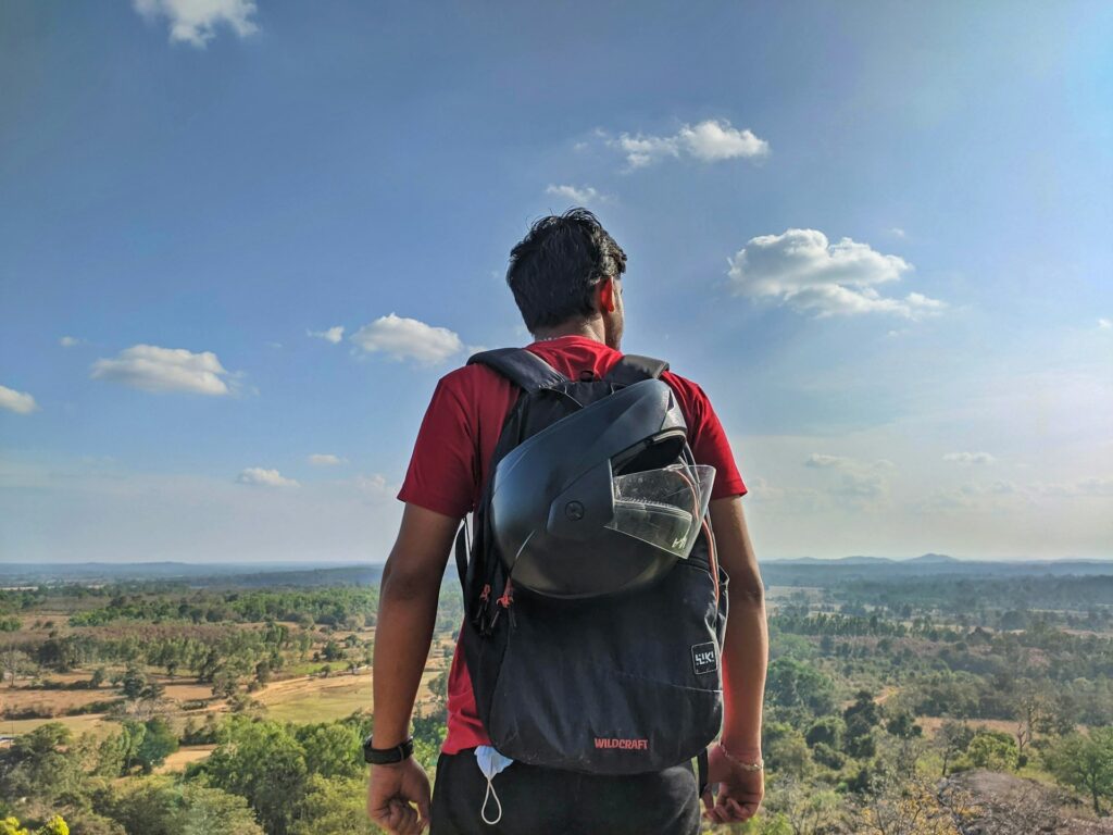 A person in a red shirt stands on a hilltop, gazing at a vast landscape and blue sky, with a backpack visible on their back.