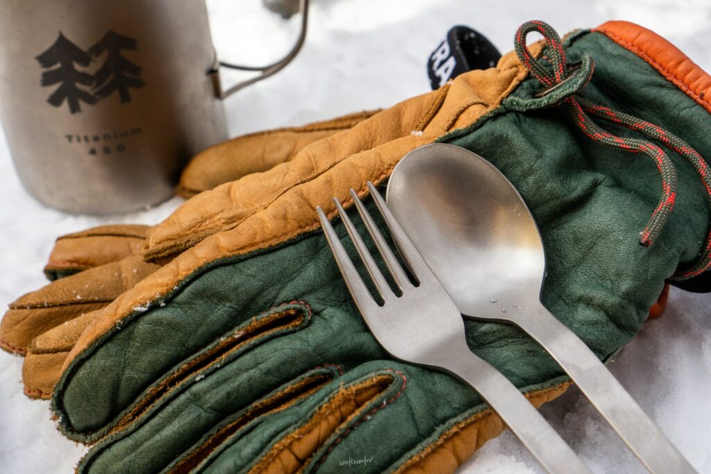 A pair of colorful gloves rests on snow, with a titanium cup in the background and a fork and spoon placed on the gloves.