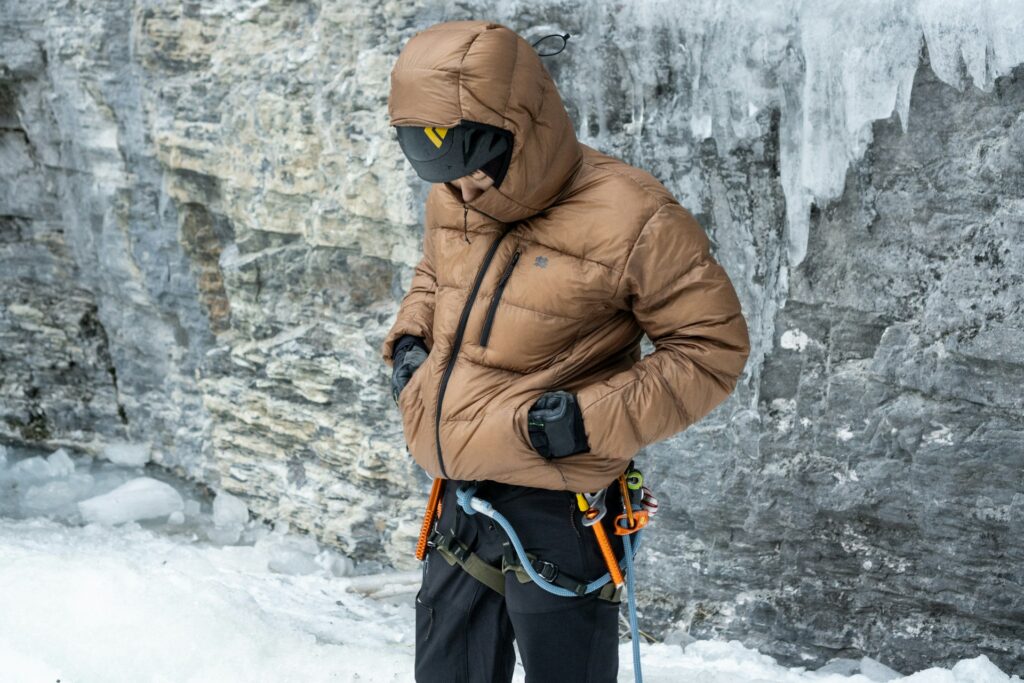 A climber in a brown puffy jacket stands on ice, adjusting gear with a helmet and gloves, against a rocky, icy backdrop.