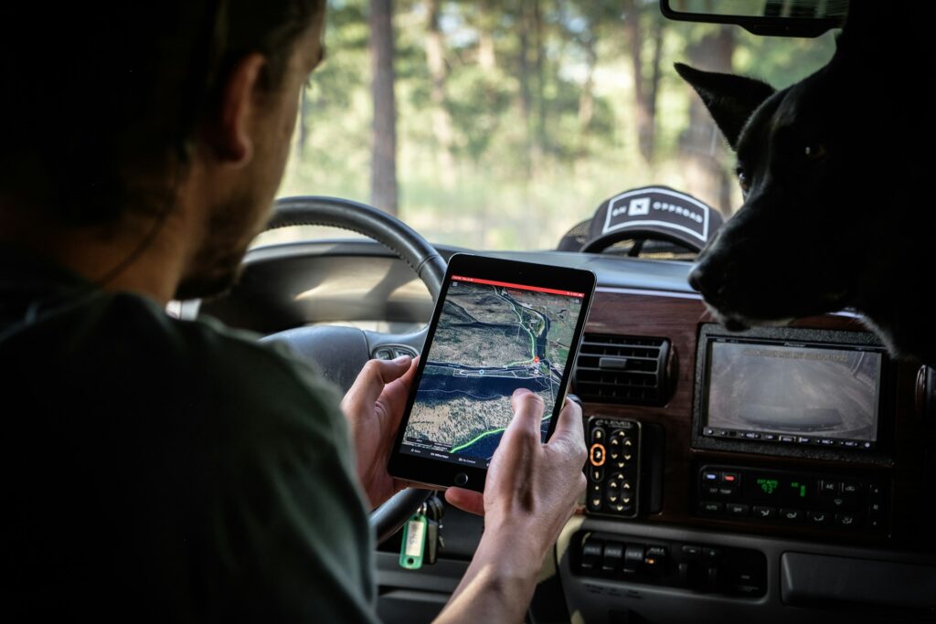 A person in a vehicle is holding a tablet displaying a detailed map, while a black dog watches from the front seat.
