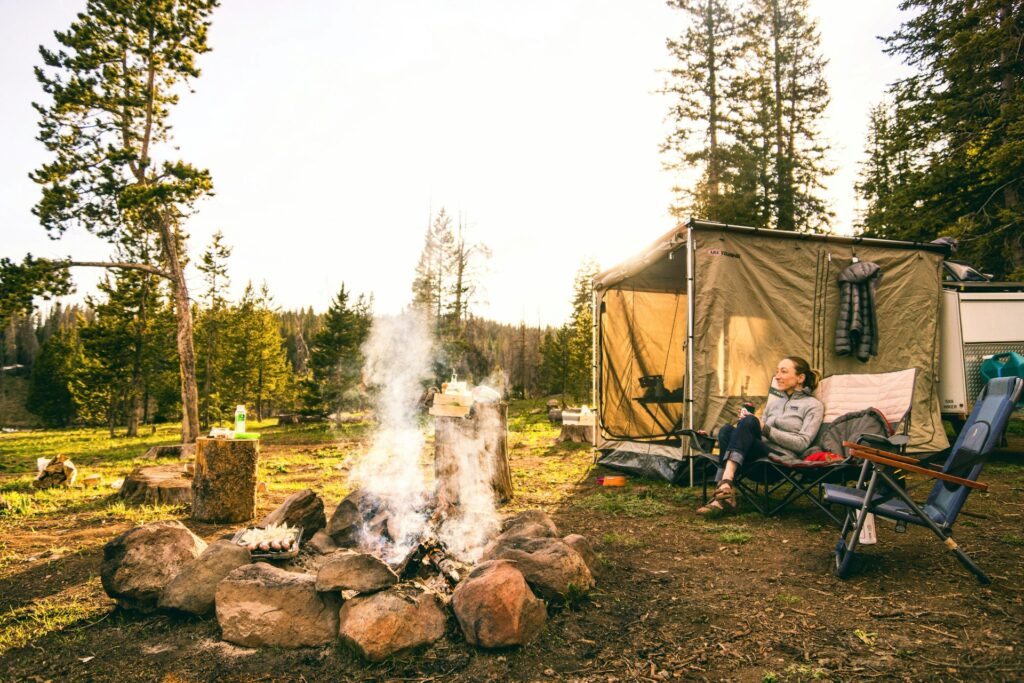 A cozy camping scene with a tent, campfire surrounded by rocks, and a person relaxing in a folding chair amidst trees and nature.