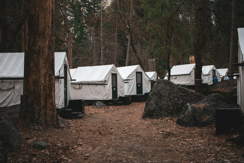 A row of white canvas tents nestled in a forest, surrounded by trees and large rocks, with a dirt path winding between them.