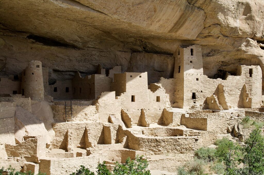 Ancient cliff dwellings with stone structures nestled under a rock overhang, surrounded by greenery.