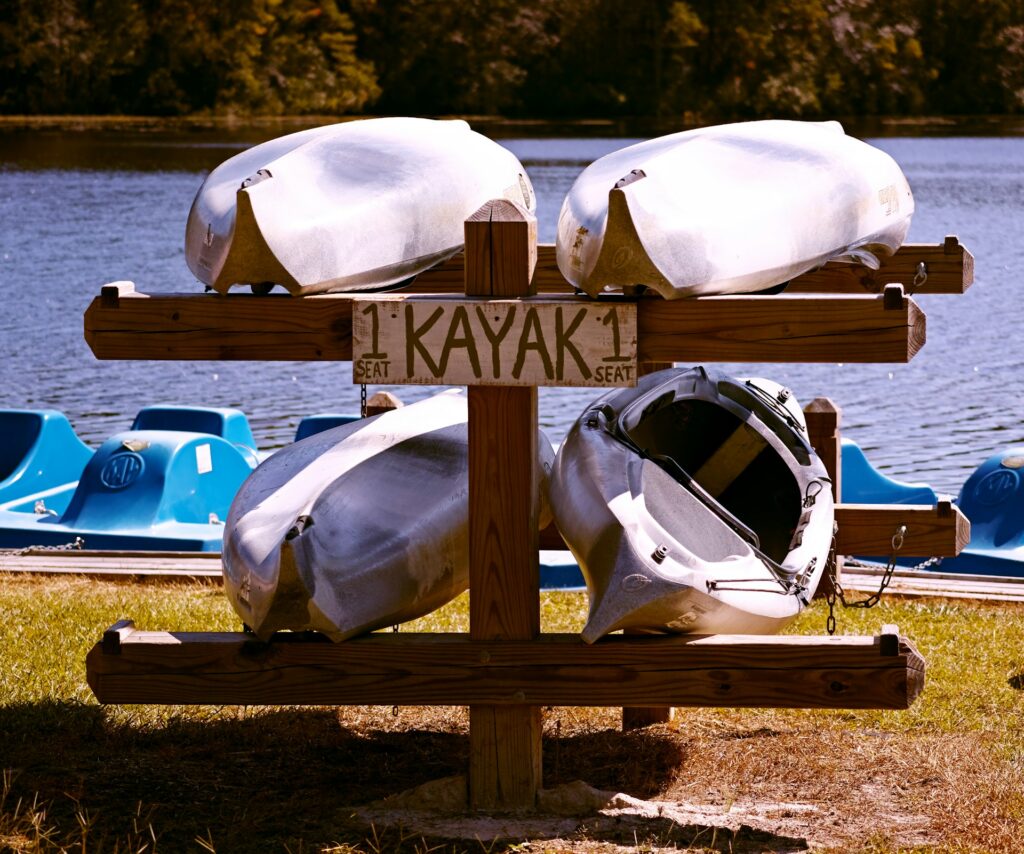 Four silver kayaks are stacked on a wooden rack labeled "1 SEAT KAYAK" beside a calm lake with blue paddle boats in the background.