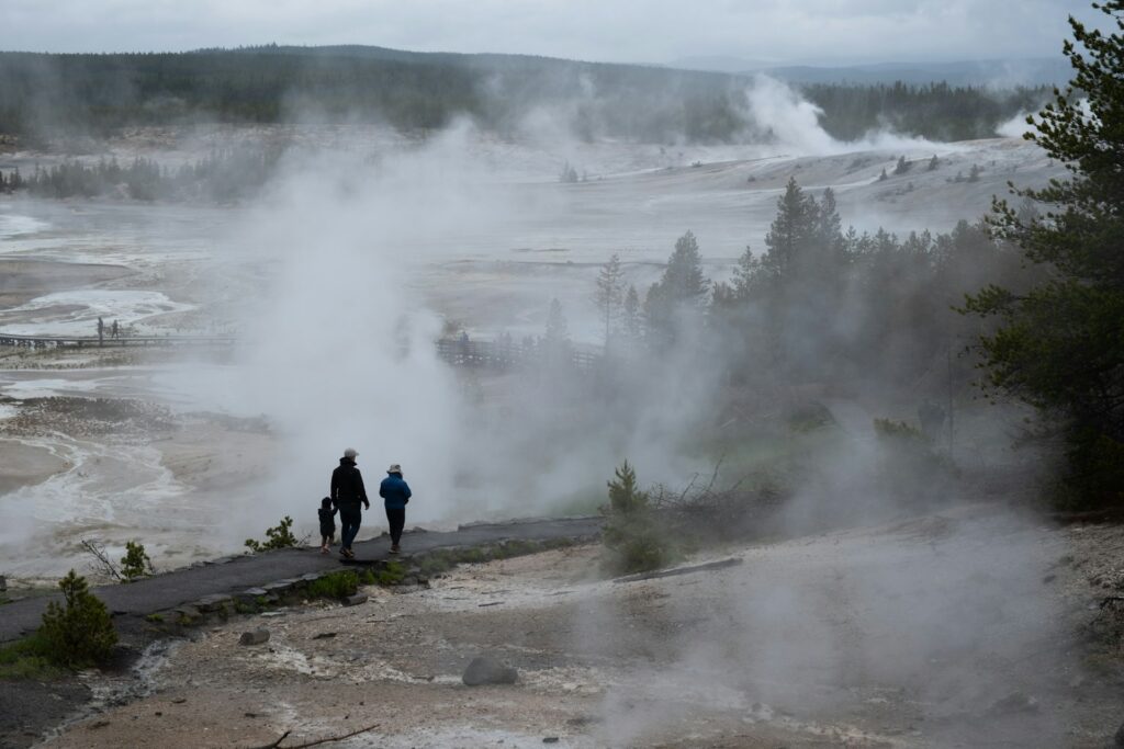 A family walks along a misty path in Yellowstone National Park, surrounded by steam and geothermal features.