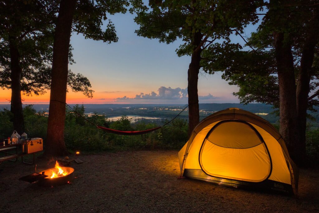 A cozy camping scene at dusk, featuring a lit tent, a campfire, a hammock, and a beautiful sunset view over the hills.