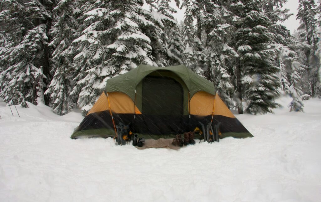 A large green and orange tent surrounded by deep snow and tall pine trees, with flakes falling in a winter wilderness setting.