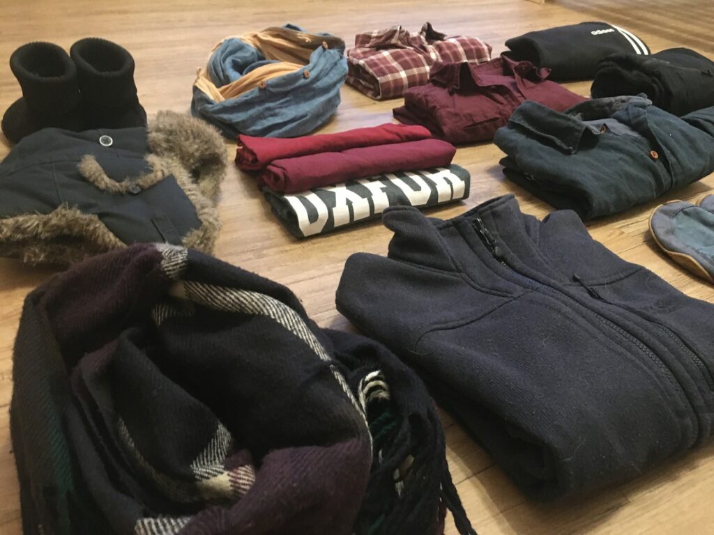 An assortment of folded winter clothing, including boots, jackets, scarves, and shirts, organized neatly on a wooden floor.
