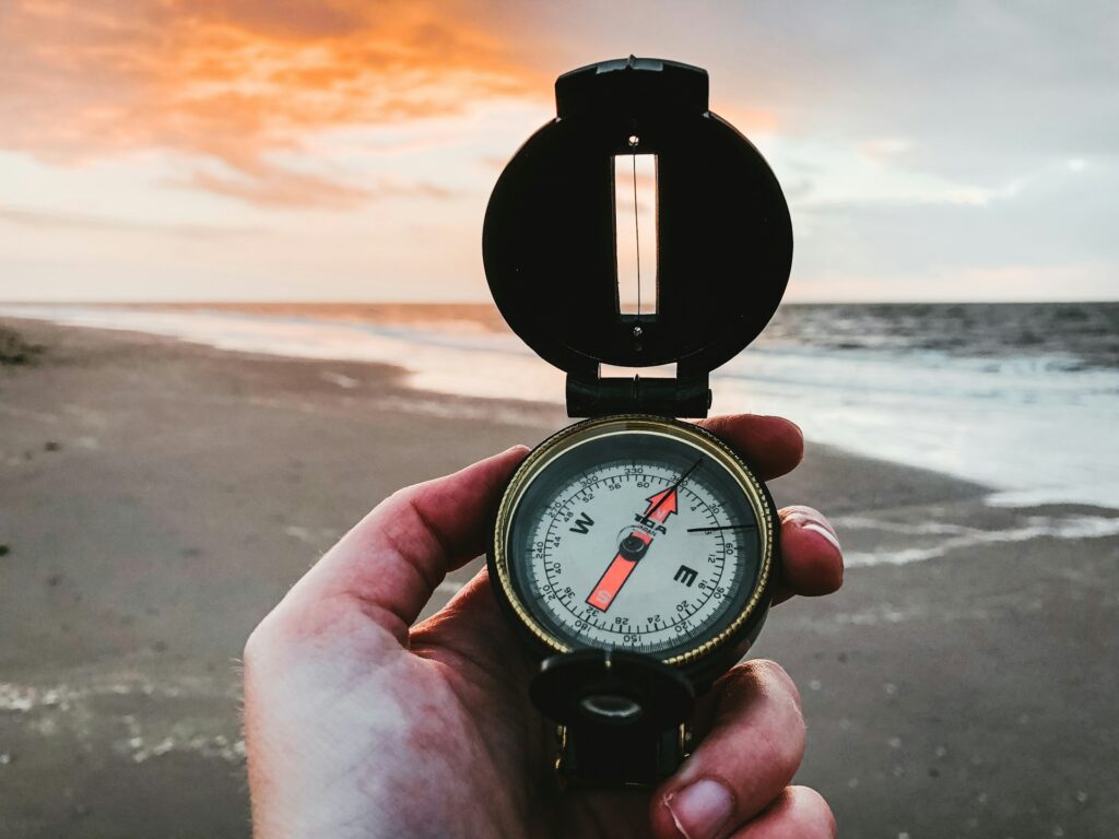 A hand holds a vintage compass at the seaside, with a vibrant sunset illuminating the sky and reflecting off the water.