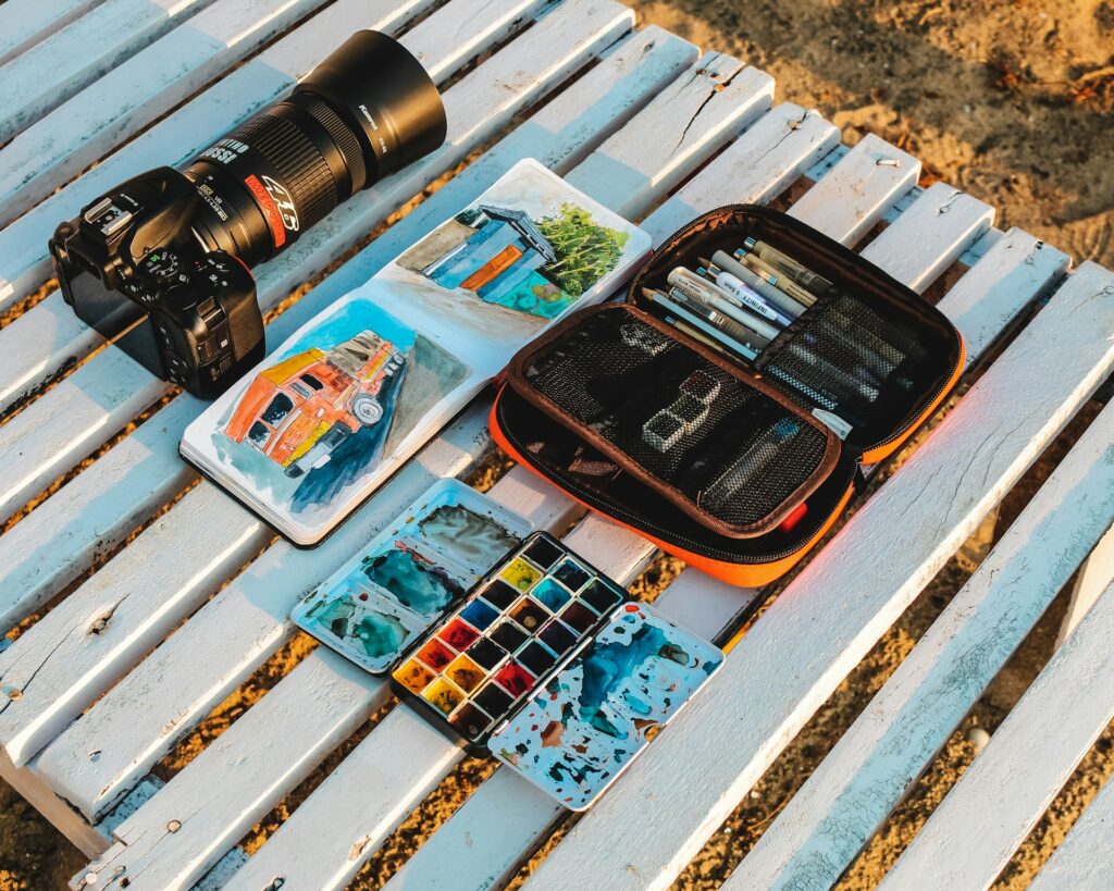 A camera, watercolor palette, sketchbook with painted truck, and a pencil case on a white wooden surface.