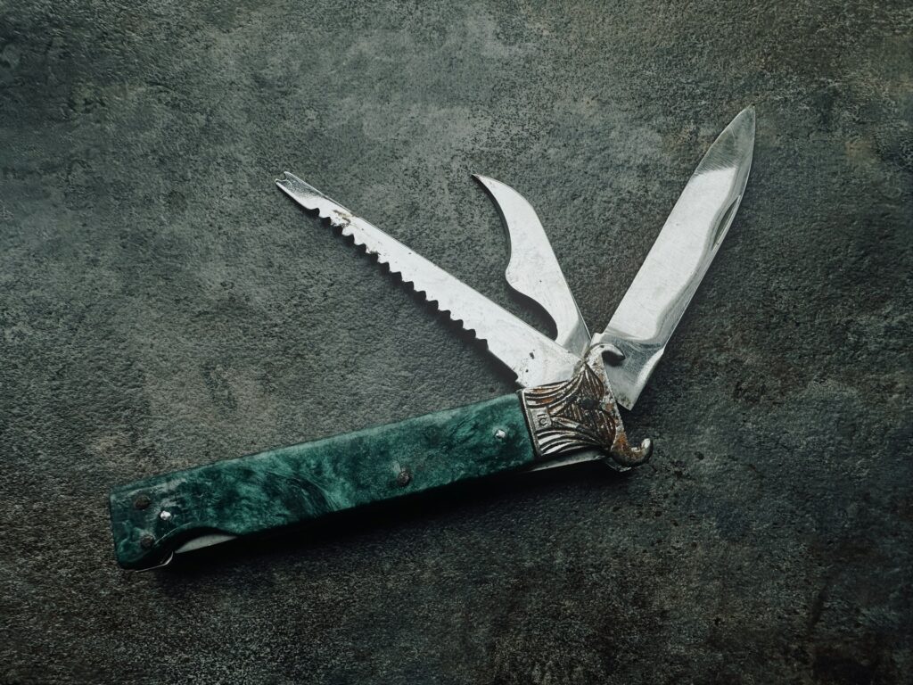 A multi-blade pocket knife with a green handle, featuring a straight edge, serrated blade, and a unique decorative metal guard.