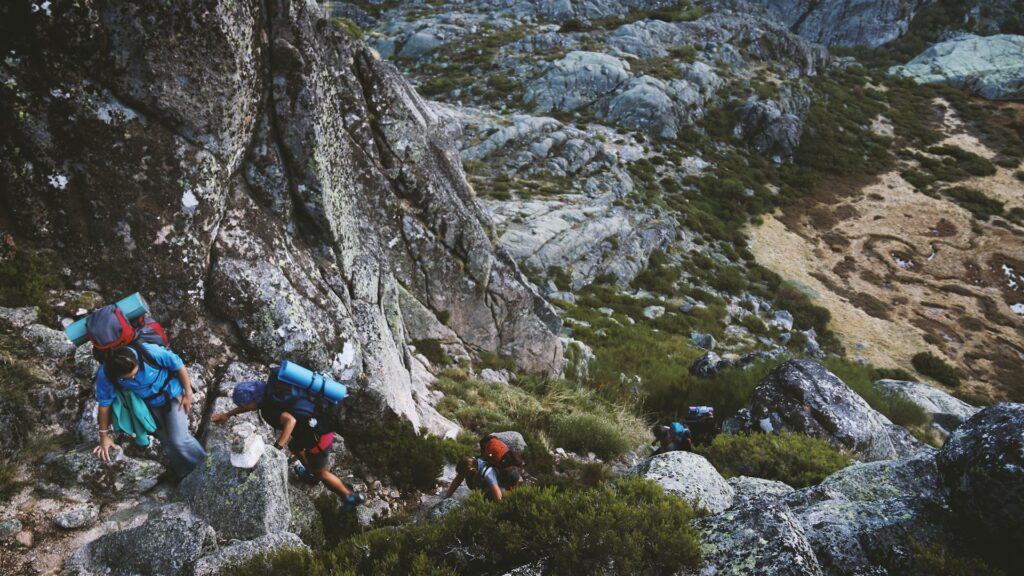 A group of hikers ascends a rocky terrain, carrying backpacks and navigating steep, uneven ground surrounded by rugged landscapes.