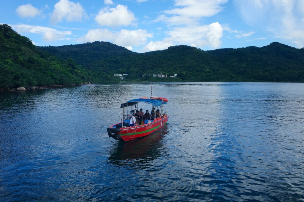A colorful boat with passengers glides through calm water, surrounded by lush green hills and a blue sky with scattered clouds.