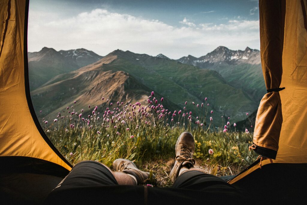 A pair of boots resting outside a tent, framed by vibrant wildflowers and stunning mountain scenery in the background.