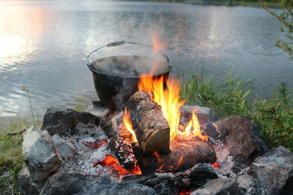 A bubbling pot over a vibrant campfire, with flames and sparks rising, set against a tranquil lakeside landscape at sunset.