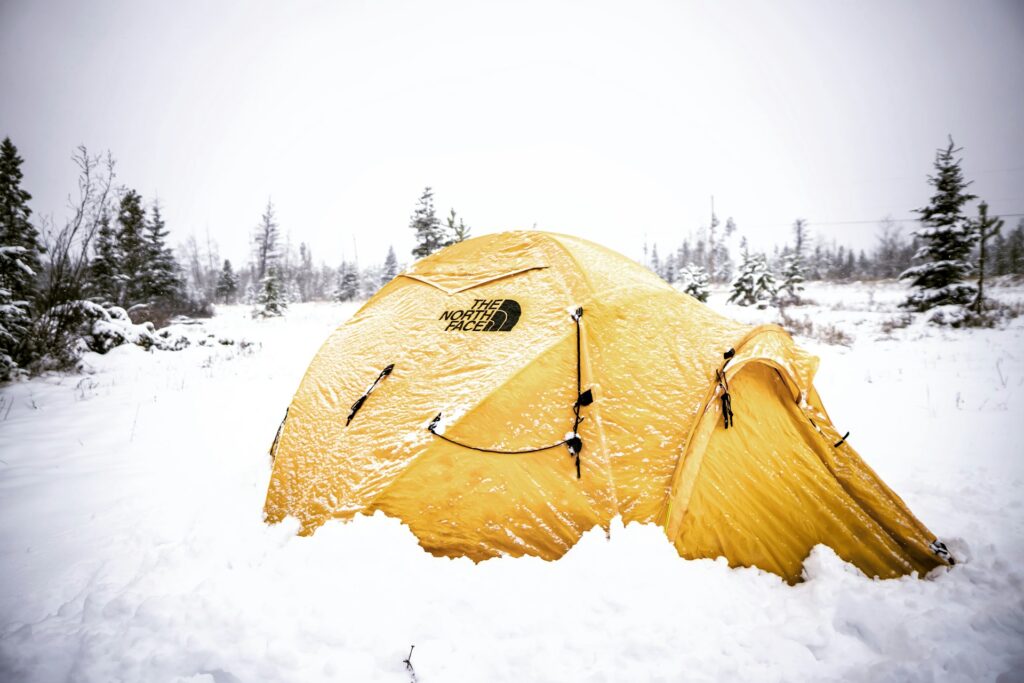 A bright yellow North Face tent partially buried in snow, surrounded by trees under a cloudy, wintry sky.