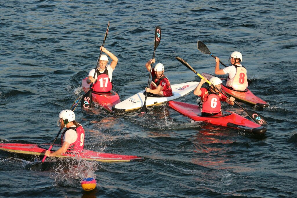A group of five kayakers in colorful boats paddle energetically across sparkling water, wearing helmets and numbered jerseys.