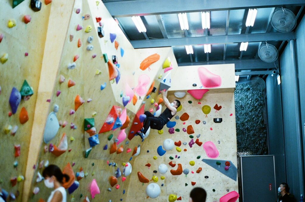 A person climbs a colorful indoor bouldering wall lined with various shaped holds, surrounded by other climbers in an active climbing gym.