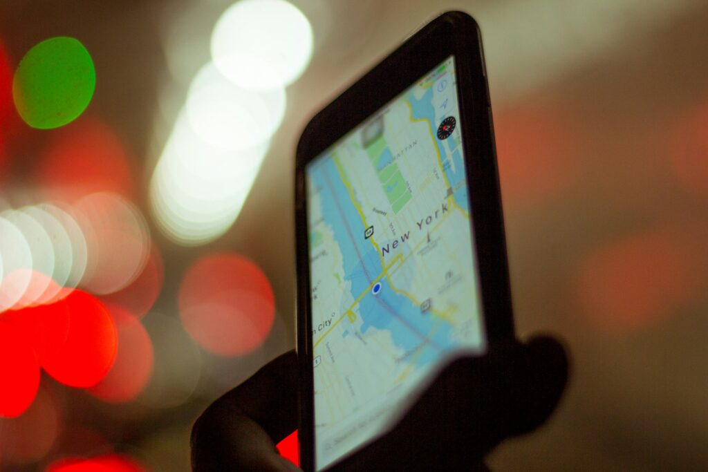 A hand holds a smartphone displaying a map of New York City, illuminated by blurred red and green lights in the background.