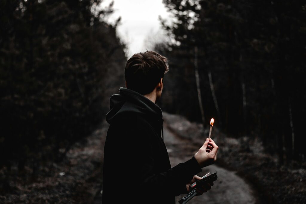 A young man stands on a dark, wooded path, holding a lit match in one hand and a lighter in the other, facing away from the camera.