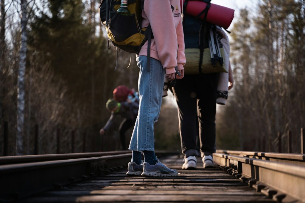 Two hikers with backpacks walk along a wooden railroad track, surrounded by trees in the background, on a bright day.