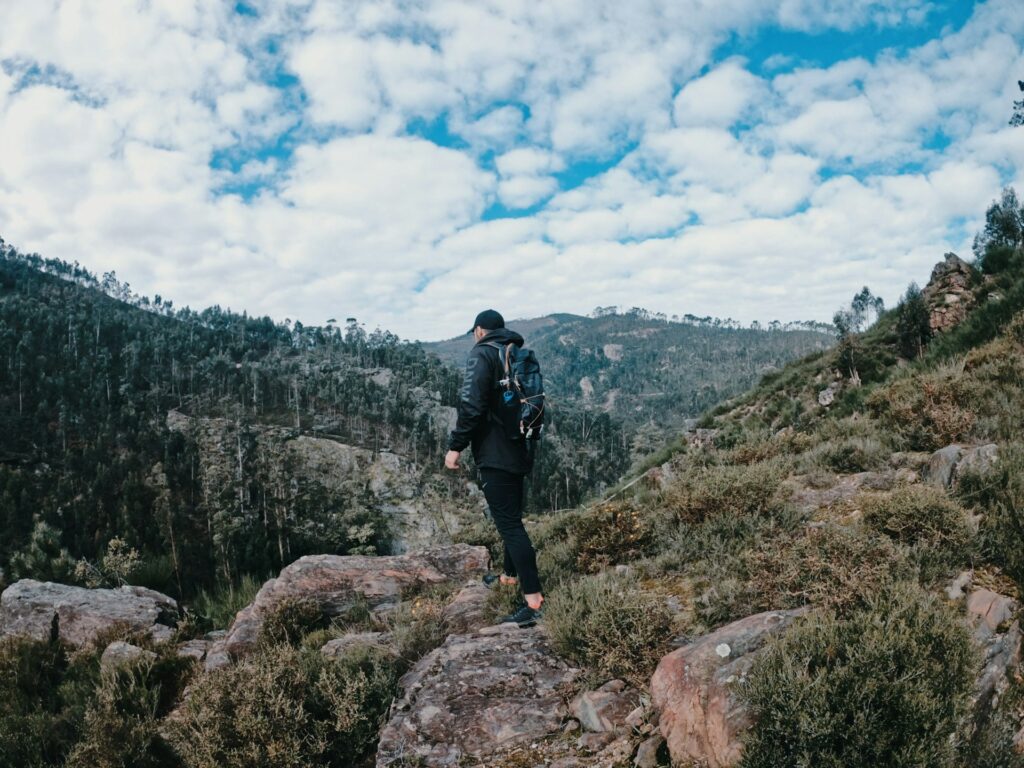 A hiker in dark clothing and a backpack walks along rocky terrain with lush greenery and a cloudy sky in the background.