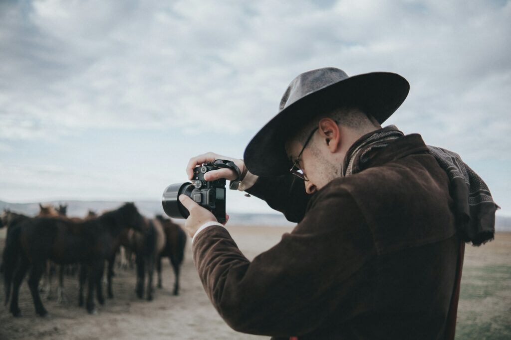 A person wearing a wide-brimmed hat takes a photograph of a group of horses in an open landscape under a cloudy sky.