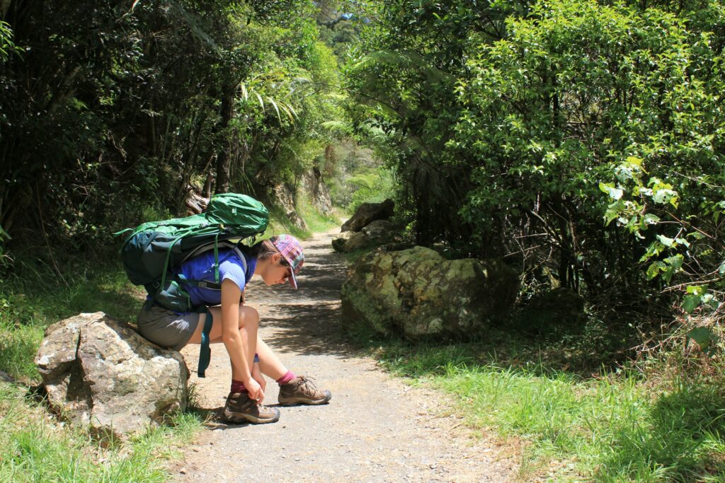 A hiker crouches on a rock, tying shoelaces beside a dirt path lined with lush greenery and natural rocks.