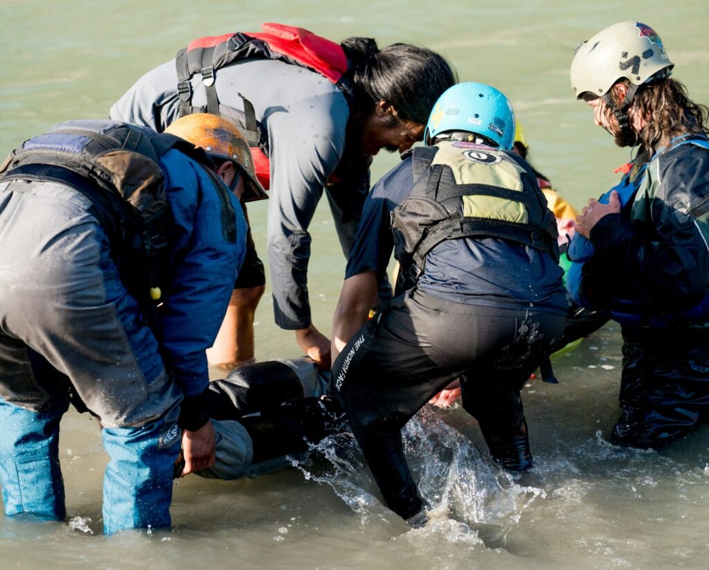 A group of six people in wet gear assists an individual in the water, splashing as they work to provide aid.