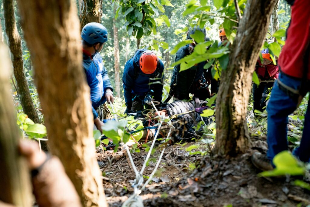 Rescue workers in helmets assist an injured person on a stretcher in a dense forest, surrounded by trees and greenery.