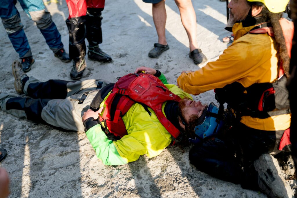 A rescue scene on a sandy beach, featuring a person in a bright yellow jacket assisting an individual lying on the ground.