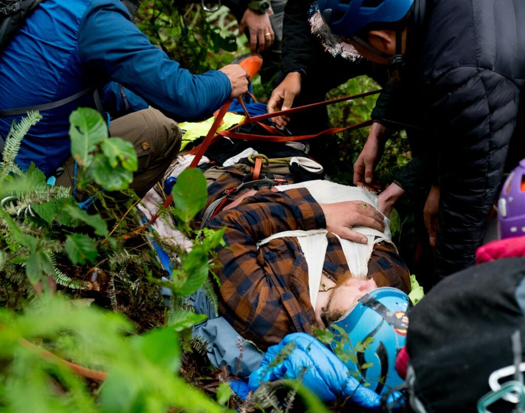 Rescuers attend to an injured climber on the forest floor, surrounded by rescue gear and teammates wearing helmets, amidst dense foliage.