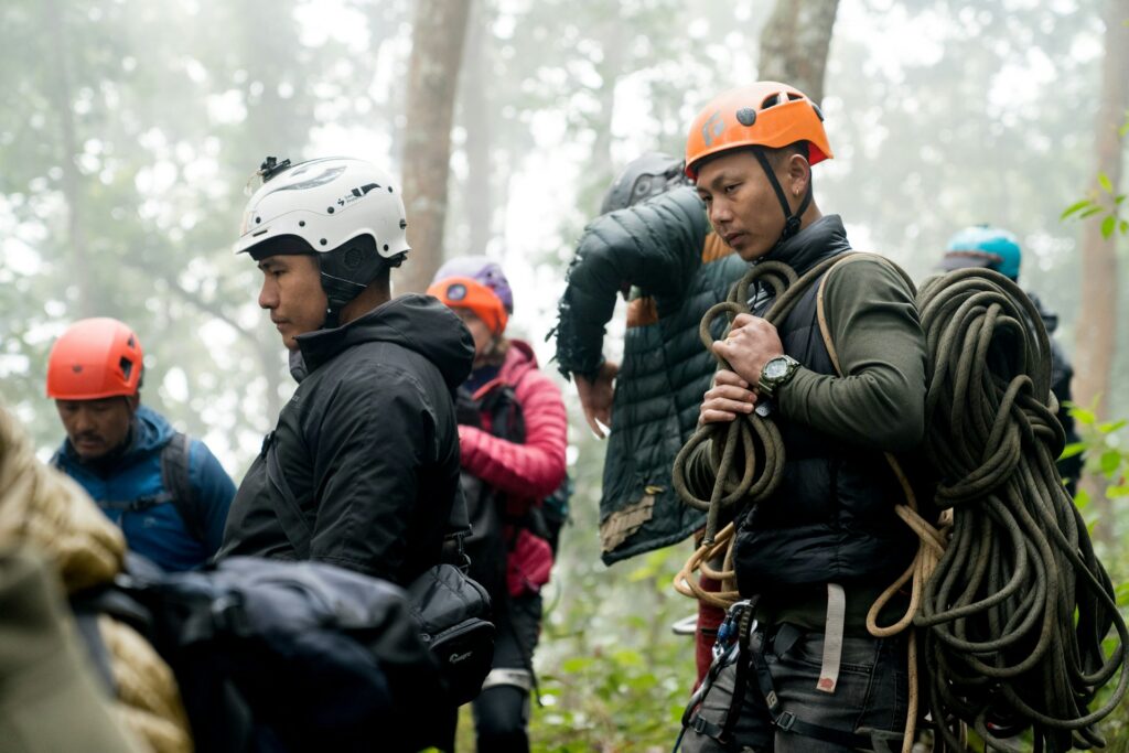 A group of climbers in helmets and gear gathers in a misty forest, preparing for an outdoor adventure, with ropes and harnesses visible.