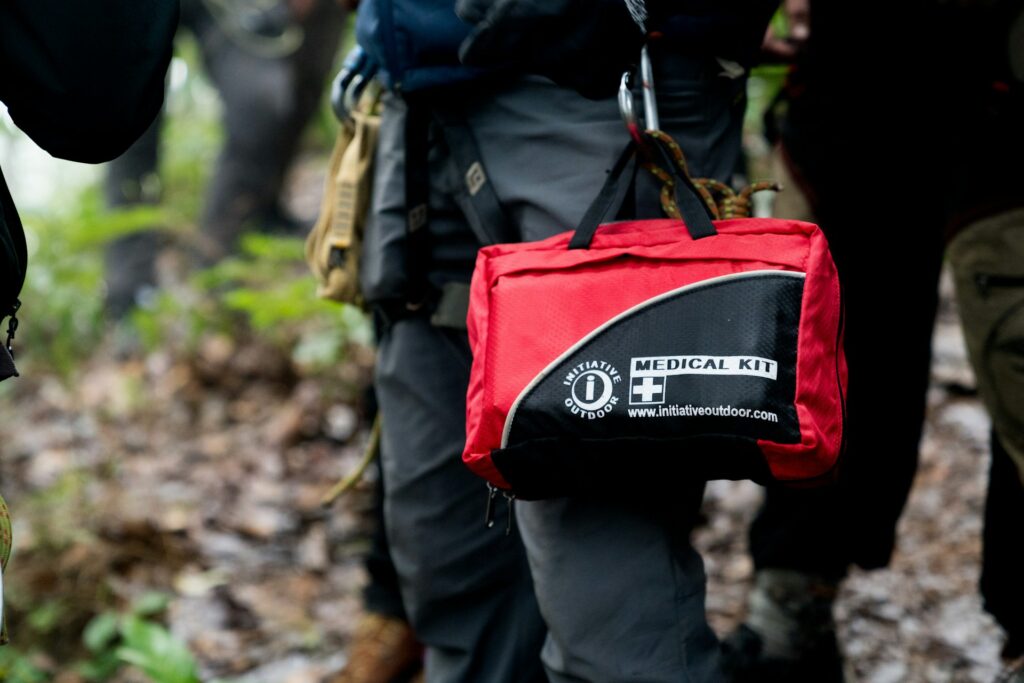 A close-up of a red medical kit labeled "INITIATIVE OUTDOOR" being held in a forest, symbolizing outdoor safety and preparedness.