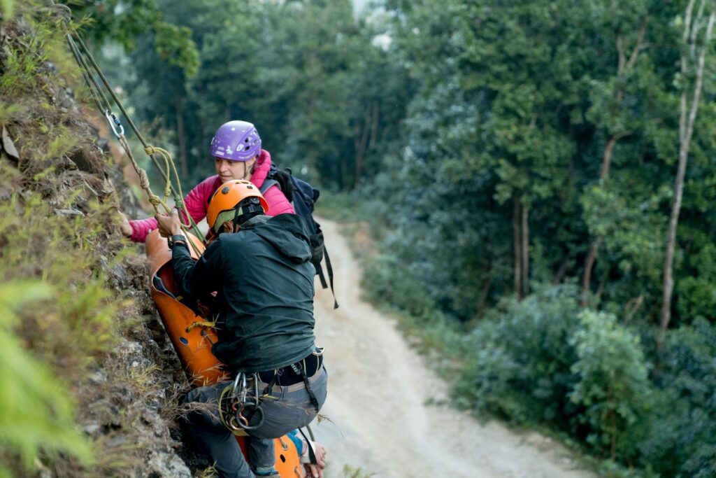 A climber in a yellow helmet assists another person in a rescue operation on a steep rock face amidst a lush green forest.