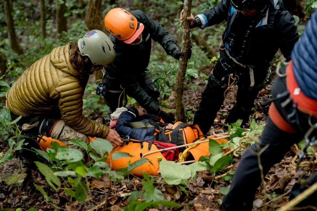 Rescue team members assist an injured climber on a steep forest slope, using a bright orange stretcher surrounded by dense foliage.