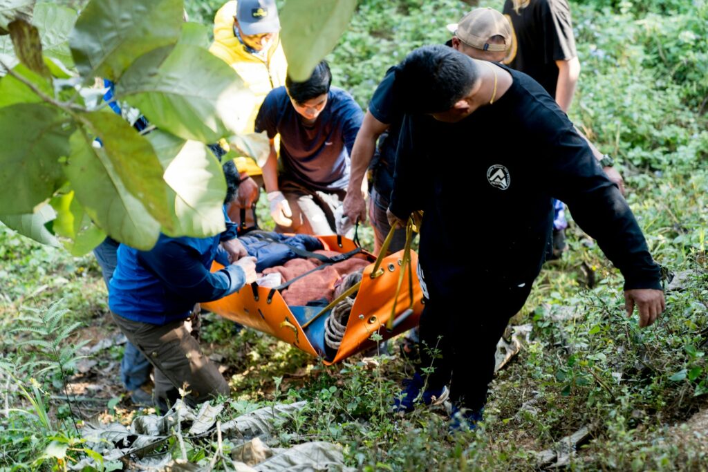 Rescue team members carry an injured person on an orange stretcher through a dense, green forested area.