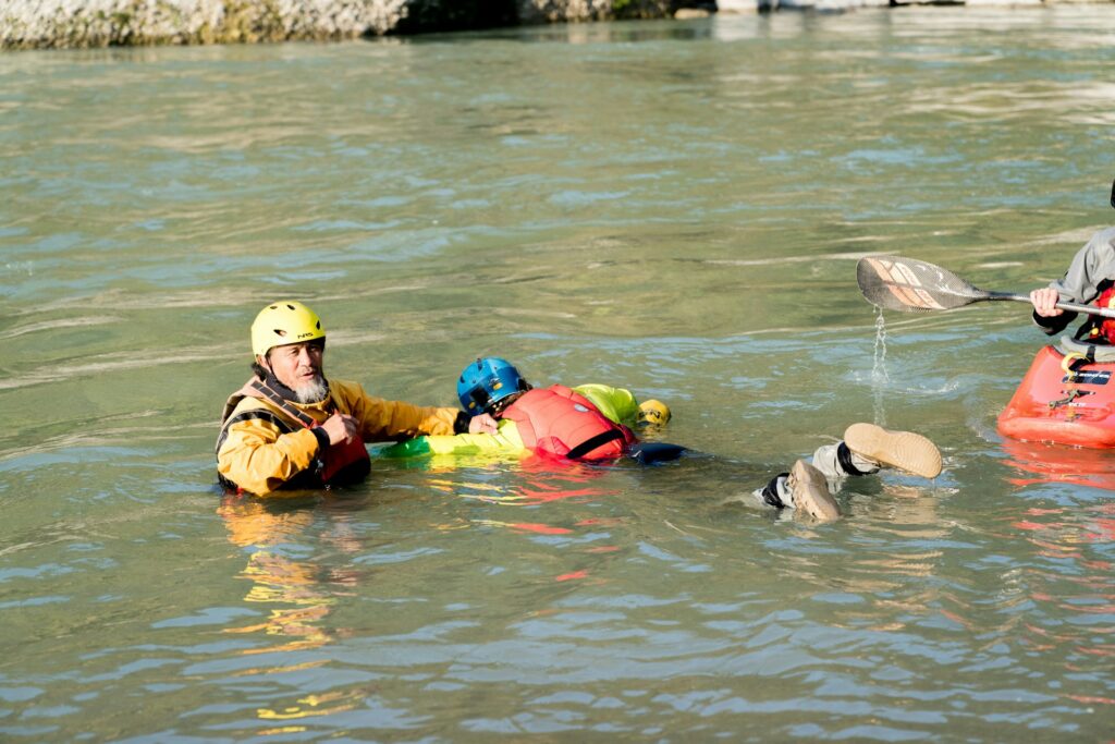 Two individuals in water rescue gear assist another person on a bright inflatable in a calm river, while a kayaker paddles nearby.
