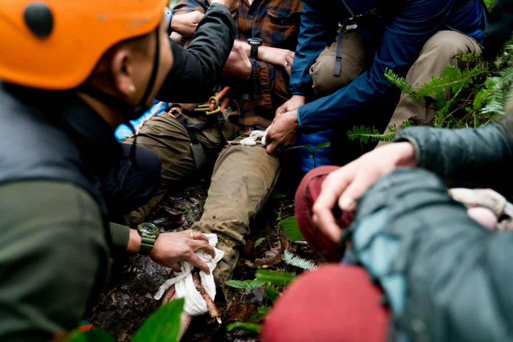 Rescue team members administer first aid to an injured person in the forest, focusing on a leg wound, surrounded by lush greenery.