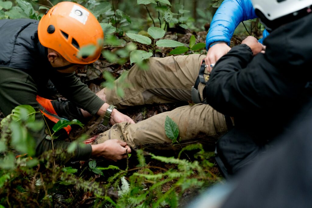Two rescuers in helmets assist an injured hiker in a dense forest, examining and tending to a leg injury with mud and foliage around.