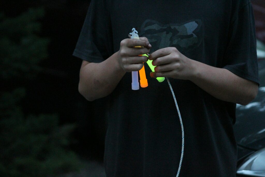 A person holds glowing glow sticks in orange, green, and purple, with a white rope, set against a dark background.